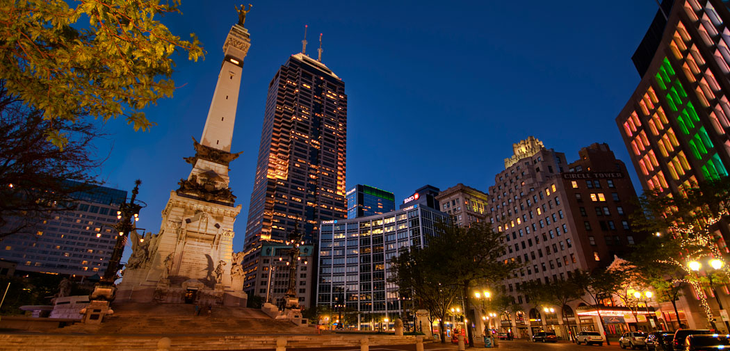 The Soldiers and Sailors Monument reaches into a blue, night sky crowded with tall buildings in Indianapolis.