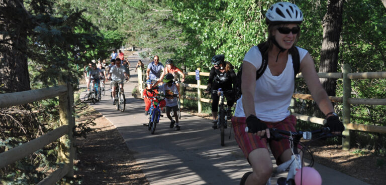 A woman leads a line of bikers, including kids along a tree covered path in Golden, CO.