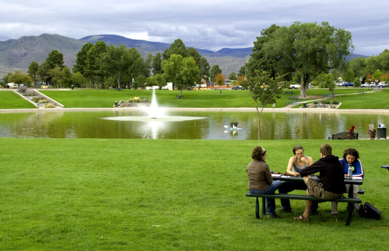A group of people city at a picnic table in front of a pond in a park that is beyond high mountains in Los Alamos.