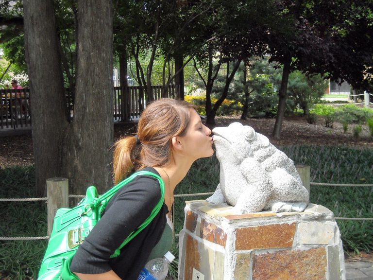 A young brown-haired woman kisses a statue of a frog at the Topeka Zoo.