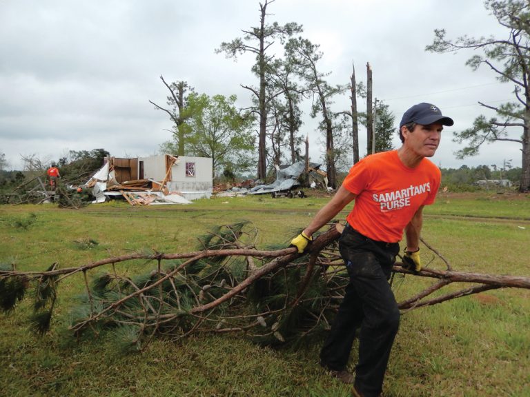 A man wearing an orange Smaritan's Purse shirt drags tree limbs away from a storm-damaged home.