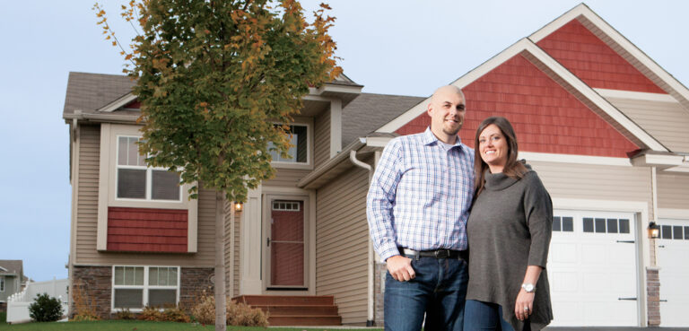 A couple stands in front of their new home in Shakopee, MN. The house has tan siding and red accents with white trim and garage doors.