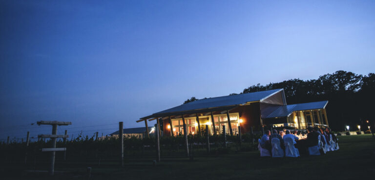 A group of people dines at a table outside at sunset at a vineyard.