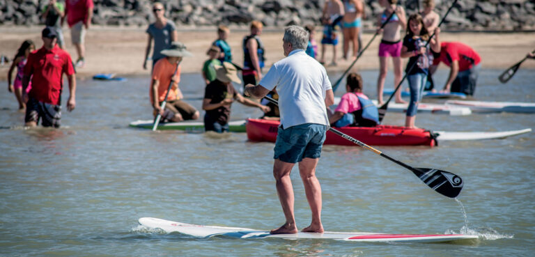 A man paddleboards at the Portneuf Wellness Complex, while many people wade in the water and watch from the beach.