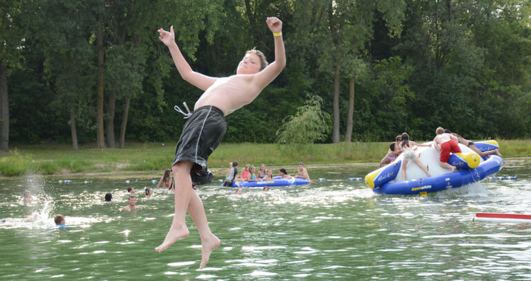 A boy falls backwards into a lake as children behind him play on an inflatable raft and other swim.