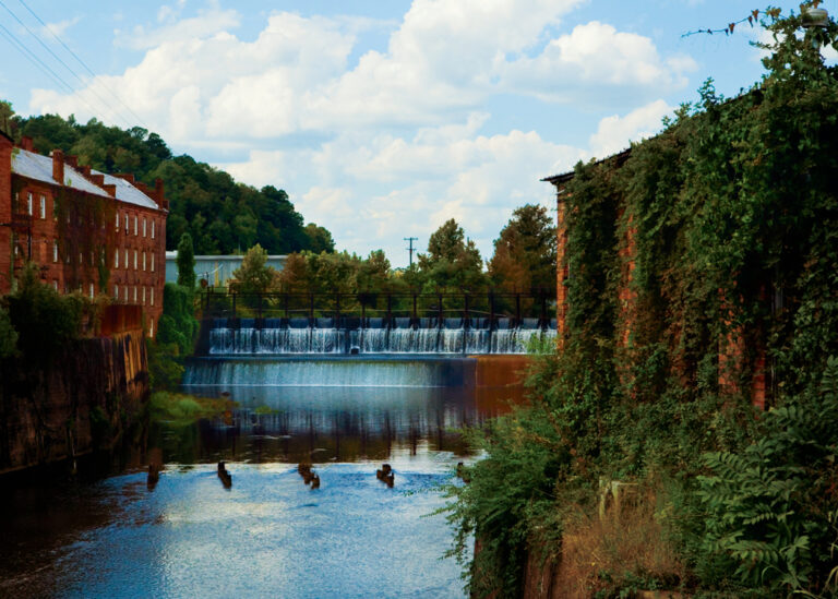 Waters from Autauga Creek flow over a dam in Prattville flanked by an old cotton gin and a brick wall covered in ivy.