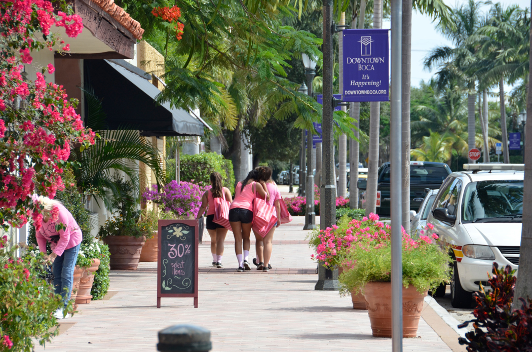 Women wearing pink tops and black shorts carry shopping bags on a brick sidewalk in downtown Boca Raton.