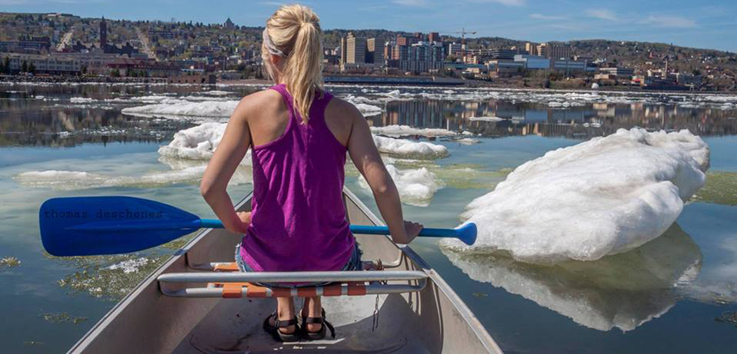 A woman canoes around huge chunks of ice in Lake Superior, overlooking the Duluth skyline.