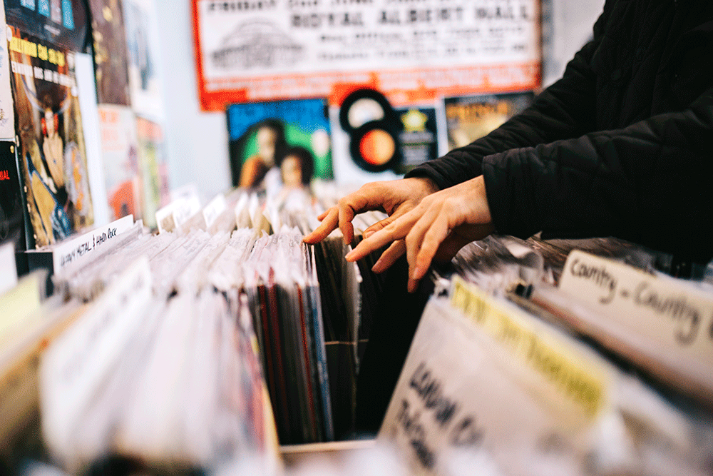 A customer searches through and selects a second hand vinyl record from a shelf in a record store, hands only, horizontal composition. Focus on hands. Close up.