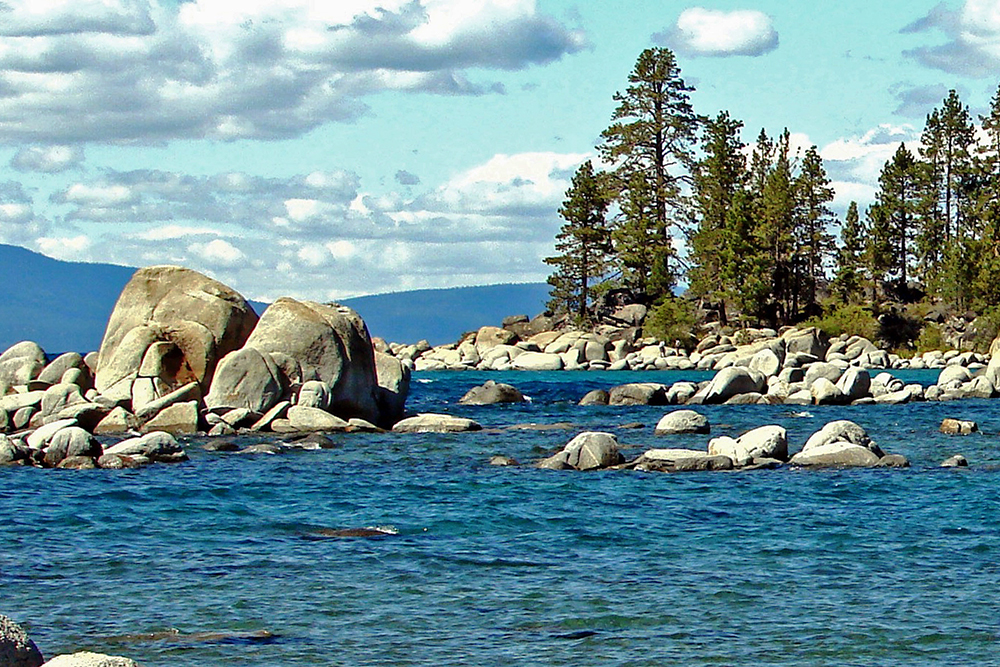 Shot of the legendary Lake Tahoe. Deep blue water, boulders, trees and the mountains in the background.