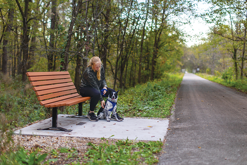 Woman sits with her dog at a bench along the Rolling Prairie Trail and Kayak, which is located in the Cedar Valley region of Iowa.
