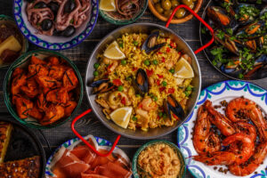 Typical spanish tapas concept. Concept include slices jamon, bowls with olives, anchovies, spicy potatoes, mashed chickpeas, shrimp, calamari, manchego with quince marmalade, pans with tortilla, paella, mussels on a wooden table.