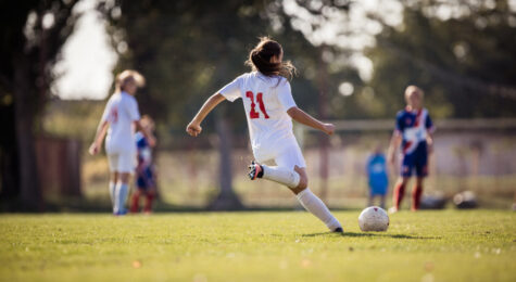 Back view of female soccer player kicking the ball during a match on a stadium. These cities support women's sports.