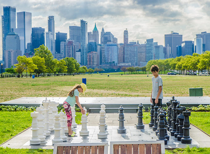 Two teenagers play chess outside on a giant chess set in Liberty State Park in Jersey City, NJ, with a view of New York City in the background. This laid-back neighborhood of NYC offers easy access to New York while boasting incredible amenities of its own.