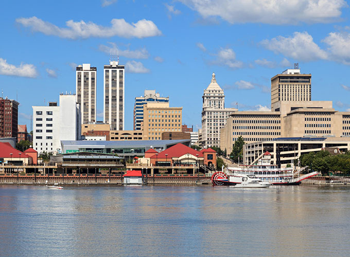 A view of Peoria, IL's riverfront against the backdrop of the downtown skyline. A manufacturing hub with an incredibly affordable housing market, Peoria is one of the best places to live in the U.S.