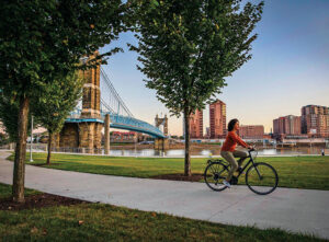 A woman rides a bicycle along the river in Cincinnati, OH. The suspension bridge in the background links the city to Covington, KY. Known as the "Queen City" for its beauty and culture, Cincinnati is ranked as one of the best places to live in the U.S. in 2023.