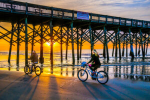 A couple ride their bicycles along the water's edge beneath the wooden pilings of the Folly Beach Pier as the morning sun slowly rises in the East.