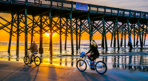 A couple ride their bicycles along the water's edge beneath the wooden pilings of the Folly Beach Pier as the morning sun slowly rises in the East.