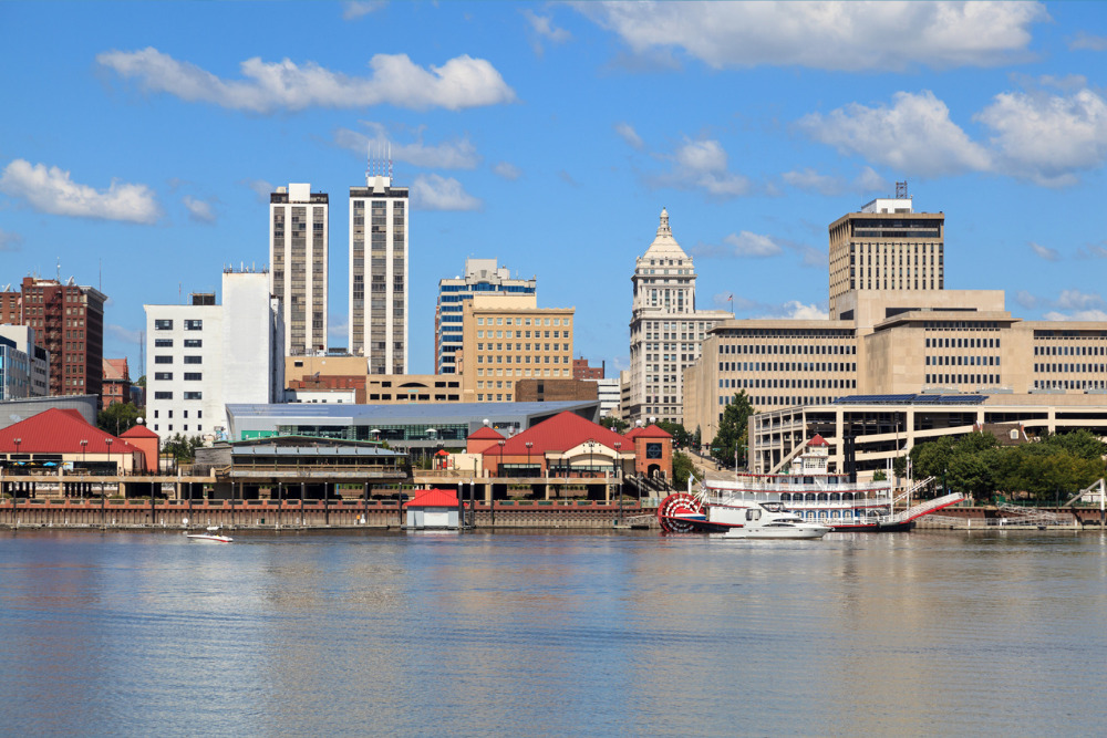 View of Peoria's river front showing downtown buildings, bridge, the Illinois river, blue sky in background.