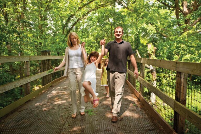 The Scotter family enjoys the Wildlife Prairie Park. The parents walk hand-in-hand with one of their daughters and swing her as they cross a bridge. The other child lingers in the background.