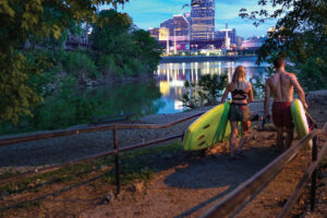 A couple carries paddleboards to the river at dusk with the downtown skyline lit up in the background.
