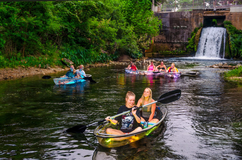 People paddle in clear kayaks in front of a waterfall in New Braunfels, TX, one of the best places to live in the U.S..