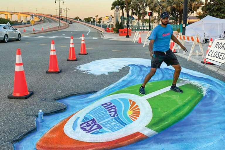 A man mimics riding a surfboard painted into a mural on the street at Daytona Beach Arts Festival.