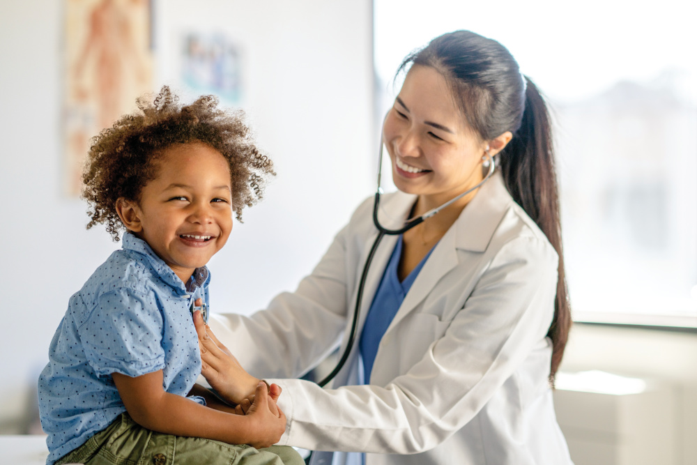 A little boy sits up on an exam table as he giggles during the physical. His female doctor is holding her stethoscope to his chest as she listens to his heart.