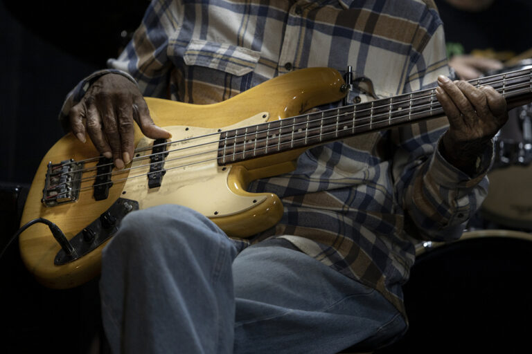 An older black man in a plaid short plays a bass guitar whle sitting in a chair.