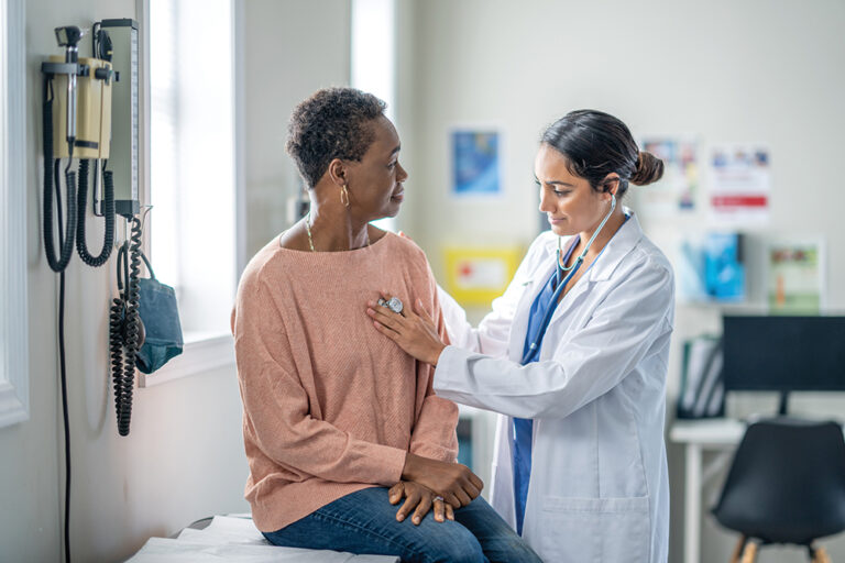A senior woman of African decent sits up on an exam table during a routine check-up. She is sitting still and quiet as her female doctor listens to her heart.