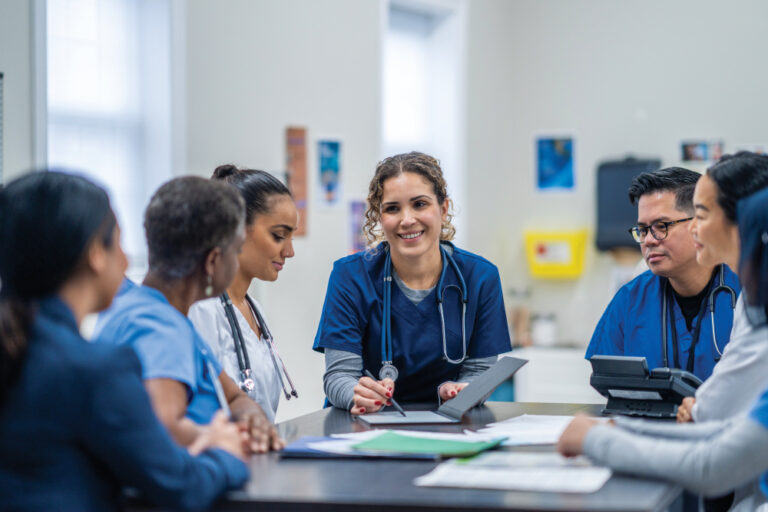 A small group of medical professionals gather around a table as they meet together to discuss patient cases. They are each dressed professionally and have files scattered between them for reference.