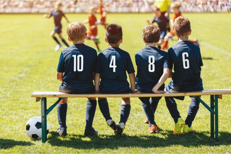 Substitute Players in Soccer Team Sitting on the Bench. Multiethnic Group of Children in Sports Team Support a Soccer Team in a Game. School Football Team Members in Blue Jerseys With Numbers stock photo