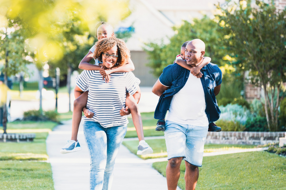 A cheerful mom and dad carry their children piggyback as they walk through their neighborhood on a sunny summer day.