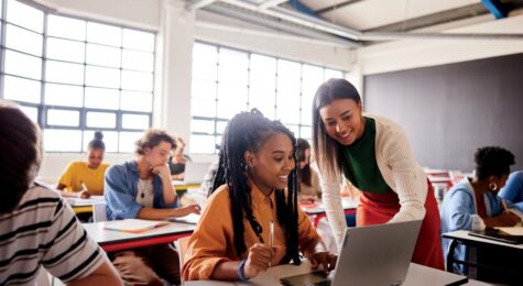 Smiling young female teacher helping a student working on a laptop during a lesson in a school classroom