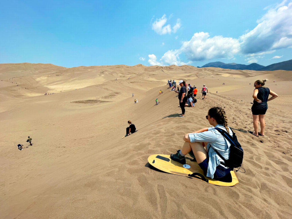 A woman sitting on a sandboard watches fellow tourists at Great Sand Dunes National Park enjoy sandboarding in Alamosa County, Colorado