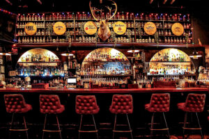 The bar at The Haberdasher in Mobile, AL, is dimly lit and features dark wood and leather seating.