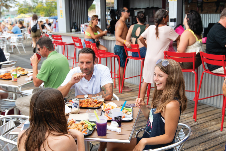 Visitors dine outdoors in the sun with views of the beach and Lake Michigan at The Deck BBQ & Beer at Pere Marquette Beach in Muskegon, Michigan. 