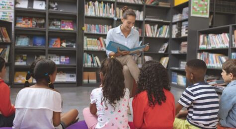 Front view of a teacher reading a story in a library to school kids sitting over big colored cushions in the foreground