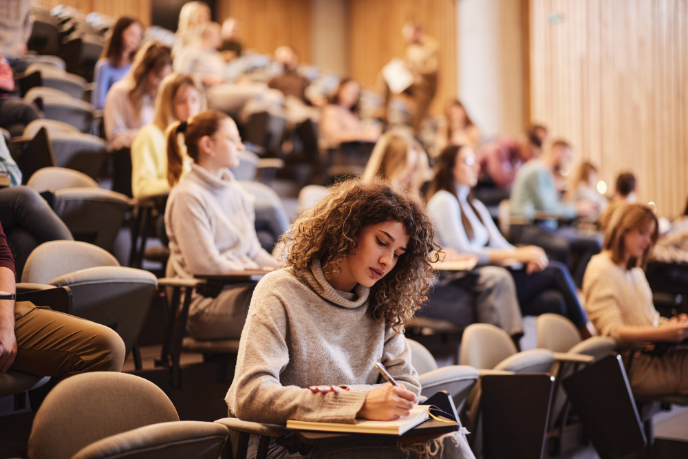 Female college student writing an exam during a at lecture hall. Her classmate are in the background.