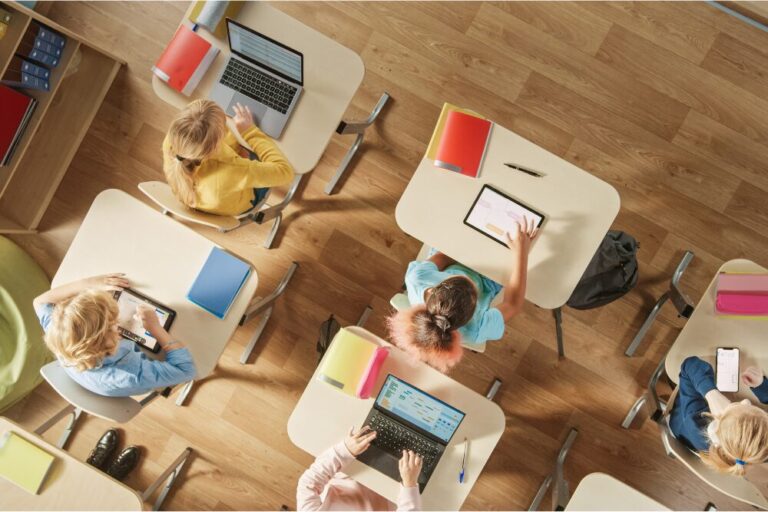 Top View Shot in Elementary School Computer Science Classroom: Children Sitting at their School Desk Using Personal Computers and Digital Tablets for Assignments.