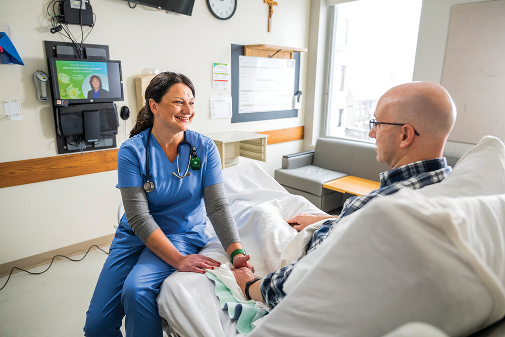 A doctor talks with an oncology patient. The doctor sits on the patient's bed and lightly holds his hand in an encouraging way.