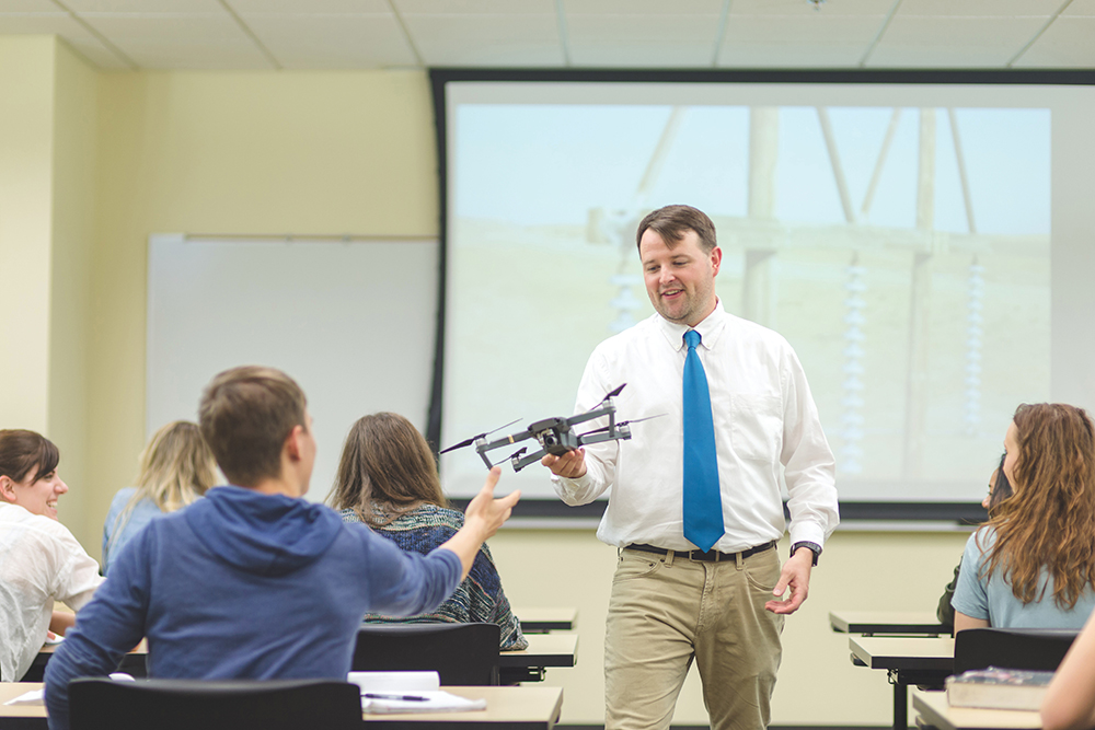 Middle-aged Caucasian male lecturing and holding drone in university classroom. The drone pilot training program at Pottsville Area High School equips students with the skills needed to fill various jobs in the drone industry.