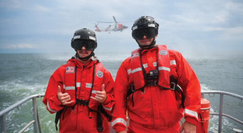 Two members from Coast Guard Station Milford Haven pose for a photo as an MH-60 Jayhawk helicopter from Coast Guard Air Station Elizabeth City hovers in the background during hoisting training in the Chesapeake Bay, Aug. 30, 2023. The crews from Coast Guard Air Station Elizabeth City conduct hoisting training weekly with various stations within the Fifth District