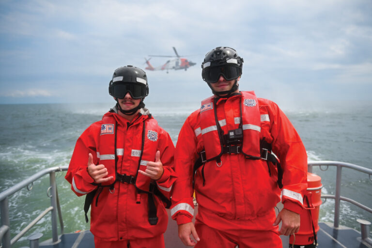 Two members from Coast Guard Station Milford Haven pose for a photo as an MH-60 Jayhawk helicopter from Coast Guard Air Station Elizabeth City hovers in the background during hoisting training in the Chesapeake Bay, Aug. 30, 2023. The crews from Coast Guard Air Station Elizabeth City conduct hoisting training weekly with various stations within the Fifth District