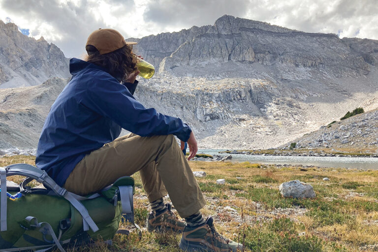 A young man sits on his backpack during a water break, against a backdrop of rocky mountains.
