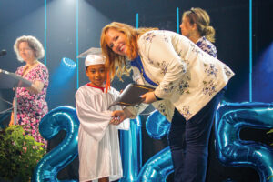 A woman shakes the hand of a young child wearing a cap and gown.