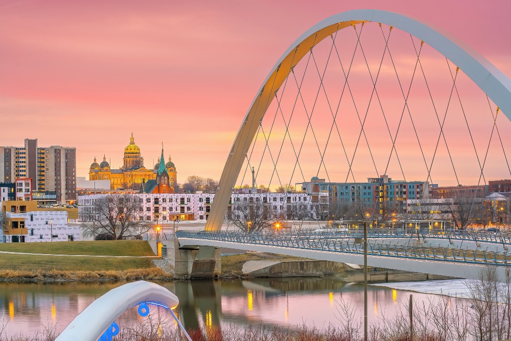 This sunset view of the downtown Des Moines city skyline features a tied arch bridge set against a pink sky.