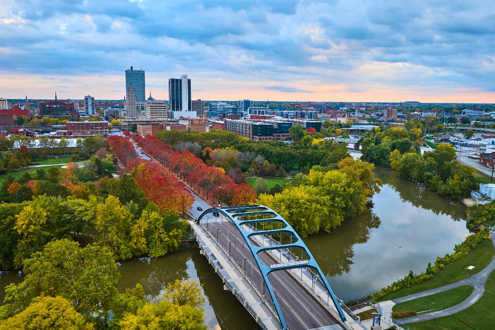 Golden hour captures autumn's vibrant hues in Fort Wayne, Indiana, featuring the modern Martin Luther King Bridge over a serene river with downtown skyline backdrop.