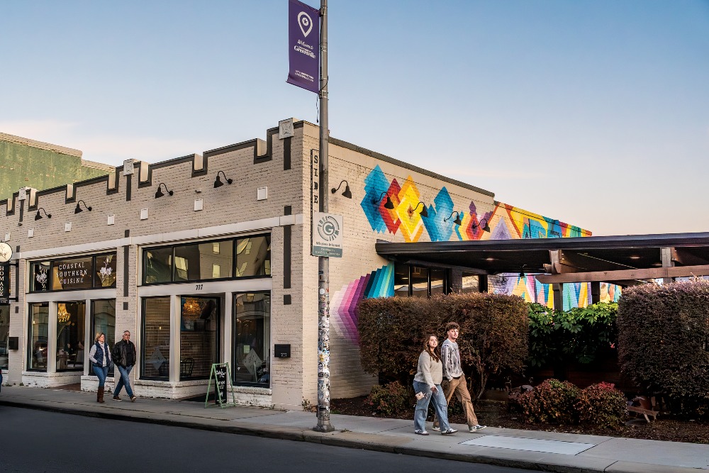 People walk through downtown Greenville, NC, against a backdrop of buildings painted with murals.