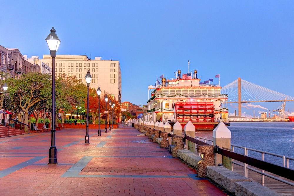 A riverboat is docked along the riverfront in Savannah, GA, at dusk against a deep blue sky.